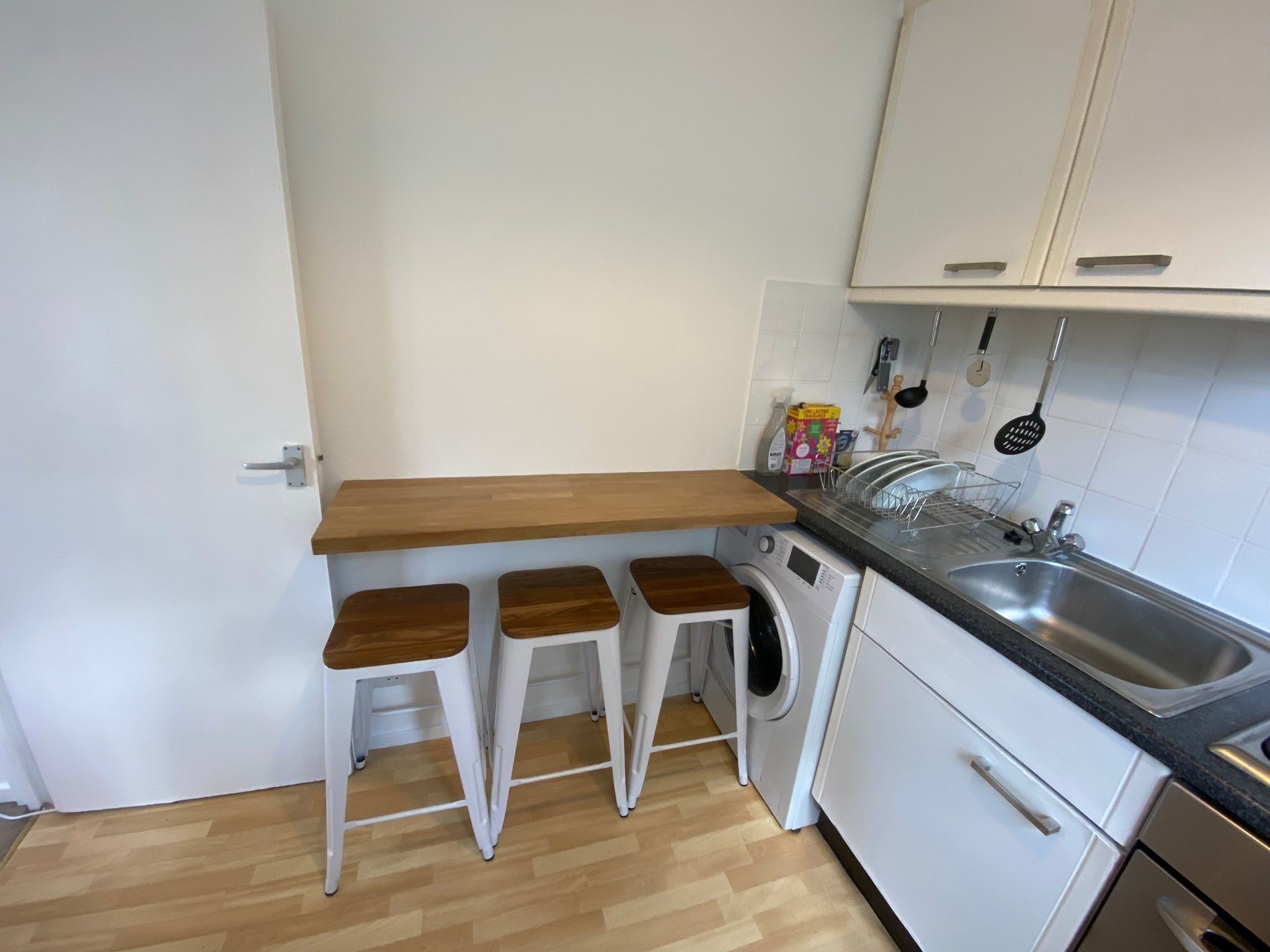 Une cuisine compacte avec un plan de travail en bois, trois tabourets blancs, un lave-linge et des placards blancs.