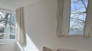 Ett modernt vardagsrum med en beige soffa, ett träbord och utsikt över kala träd genom ett fönster med gardiner.