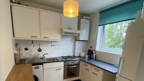 Ett kompakt kök med vita skåp, en inbyggd tvättmaskin, en gasspis och ett fönster med utsikt över grönområden.