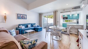 A modern living room with a blue sofa, a dining area with white chairs, and a kitchen with teal cabinets. - Relaxing Beach House in Alfamar by LovelyStay (Olhos de Água)