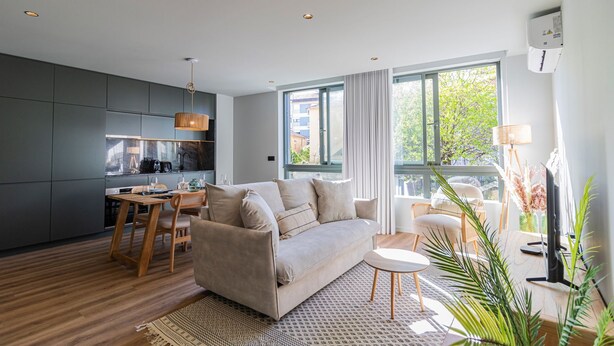 En moderne stue med sofa, spisebord og stole, et køkken med mørke skabe, samt store vinduer der giver udsigt til det fri.