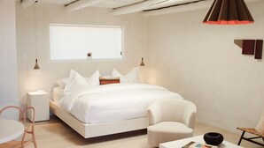 A minimalist bedroom with a bed, bedside tables, a chair, a small table, and a window. - Palace Modern Residences at the Railyard - Studio (Santa Fe)