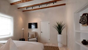 A bedroom with a bed, a chair, a TV, a door, a plant, and a shelf with decorative items.