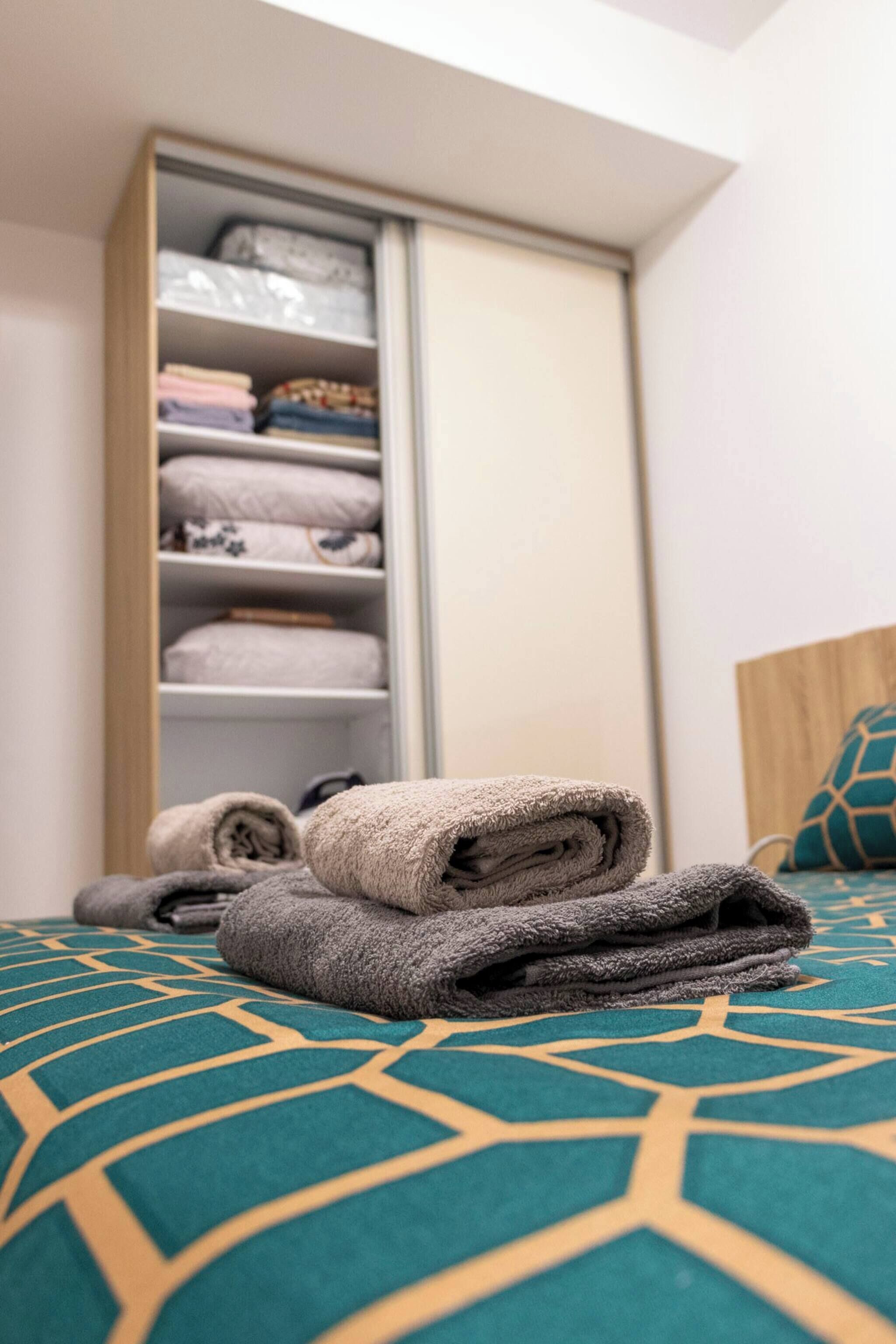 Un lit bien fait, recouvert d’une couvre-lit à motifs géométriques, avec une tête de lit en bois et un placard contenant des serviettes pliées.