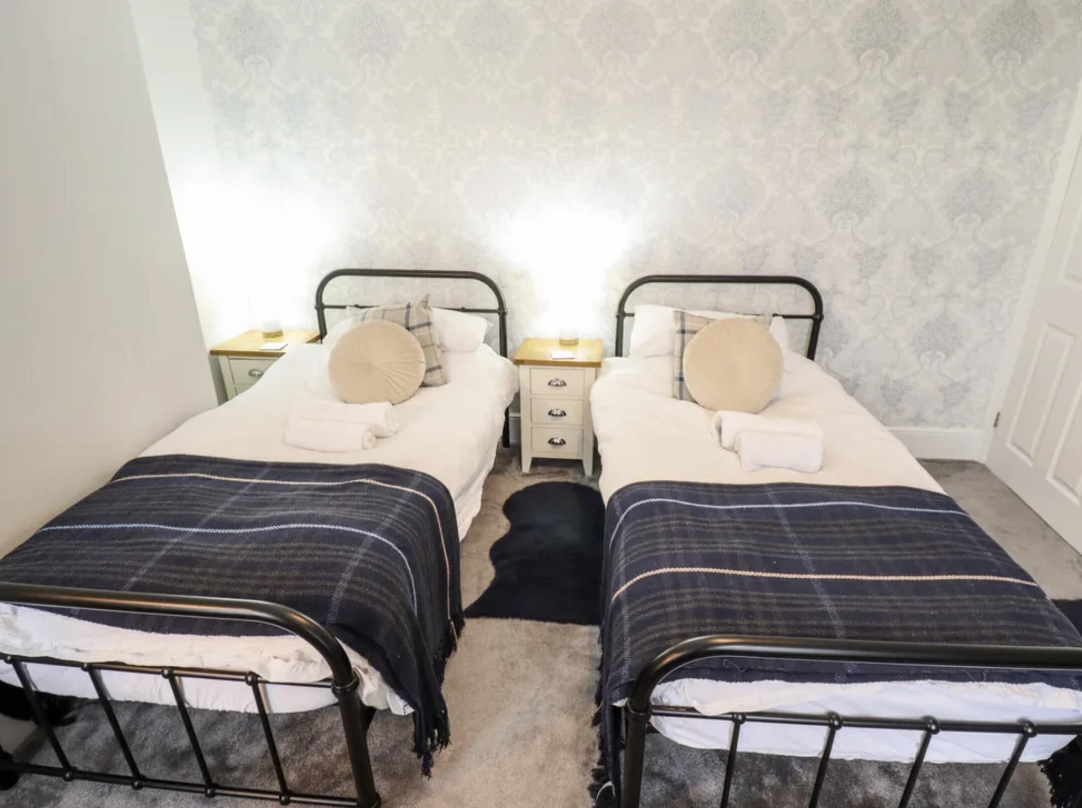 Una habitación con dos camas individuales, cada una con estructura metálica, ropa de cama blanca y una manta a rayas oscuras. Hay mesitas de noche con lámparas y un papel tapiz estampado en las paredes.