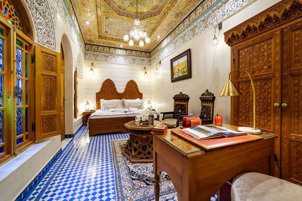 Quarto com piso xadrez, cama com cabeceira de madeira, escrivaninha com luminária e portas de madeira ornamentadas.