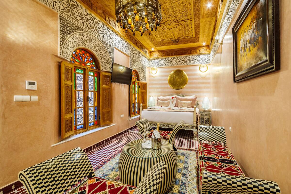 Quarto com detalhes elaborados nas paredes e no teto, uma cama com almofadas, uma televisão e um banquinho estampado.
