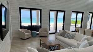 A spacious living area with a large window overlooking a beach, a sofa, a coffee table, and a side table. - Steps From the Water on One of The Most Beautiful Beaches in Exuma! (Rokers Point Settlement)