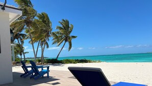A beachside lounge with blue chairs and a wooden deck, overlooking a turquoise ocean and palm trees. - Steps From the Water on One of The Most Beautiful Beaches in Exuma! (Rokers Point Settlement)