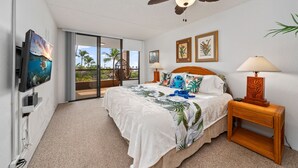 Ein Schlafzimmer mit Bett, Deckenventilator, Fernseher und Blick auf Palmen durch eine Schiebetür.