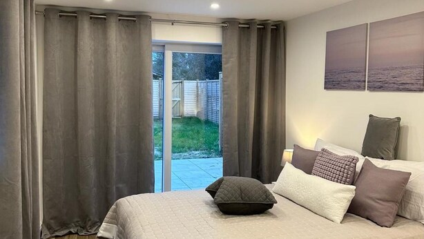 Une chambre à coucher avec un grand lit, des rideaux gris, une vue sur l’extérieur et un tableau accroché au mur.