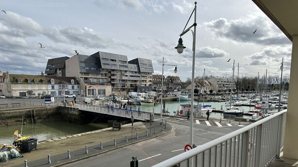 A marina with boats, a pedestrian bridge, and buildings in the background. - Appartement Le Royal vue Port (Courseulles-sur-Mer)