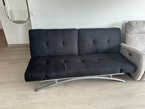 A black sofa with a metal frame, a light-colored armchair, and a white wall with a light switch. - Appartement Le Royal vue Port (Courseulles-sur-Mer)