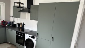 A compact kitchen with grey cabinets, a built-in washing machine, and a coffee maker on the counter. - Appartement Le Royal vue Port (Courseulles-sur-Mer)