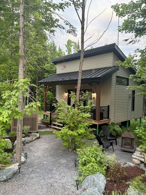 A two-story cabin with a covered porch, surrounded by a gravel driveway and a wooded area. - Tiny cottage for 2 -SPA & outdoor -Le EVA (Adstock)
