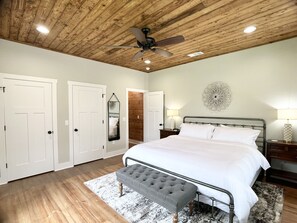 Un dormitorio con techo de madera, una cama con ropa de cama blanca, un banco gris y un elemento decorativo en la pared.