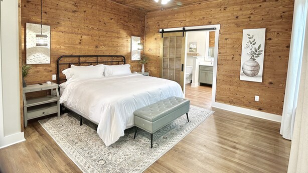 A bedroom with a wooden ceiling, a bed, a nightstand, a bench, and a door leading to a bathroom. - Luxury Home Near Crater of Diamonds 
3 King Beds 2 Full Baths
Sleeps 7 (Nashville)