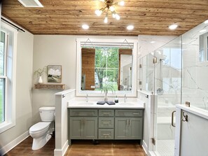 Un baño moderno con techo de madera, un ventanal amplio, un lavamanos blanco y un box de ducha de vidrio.