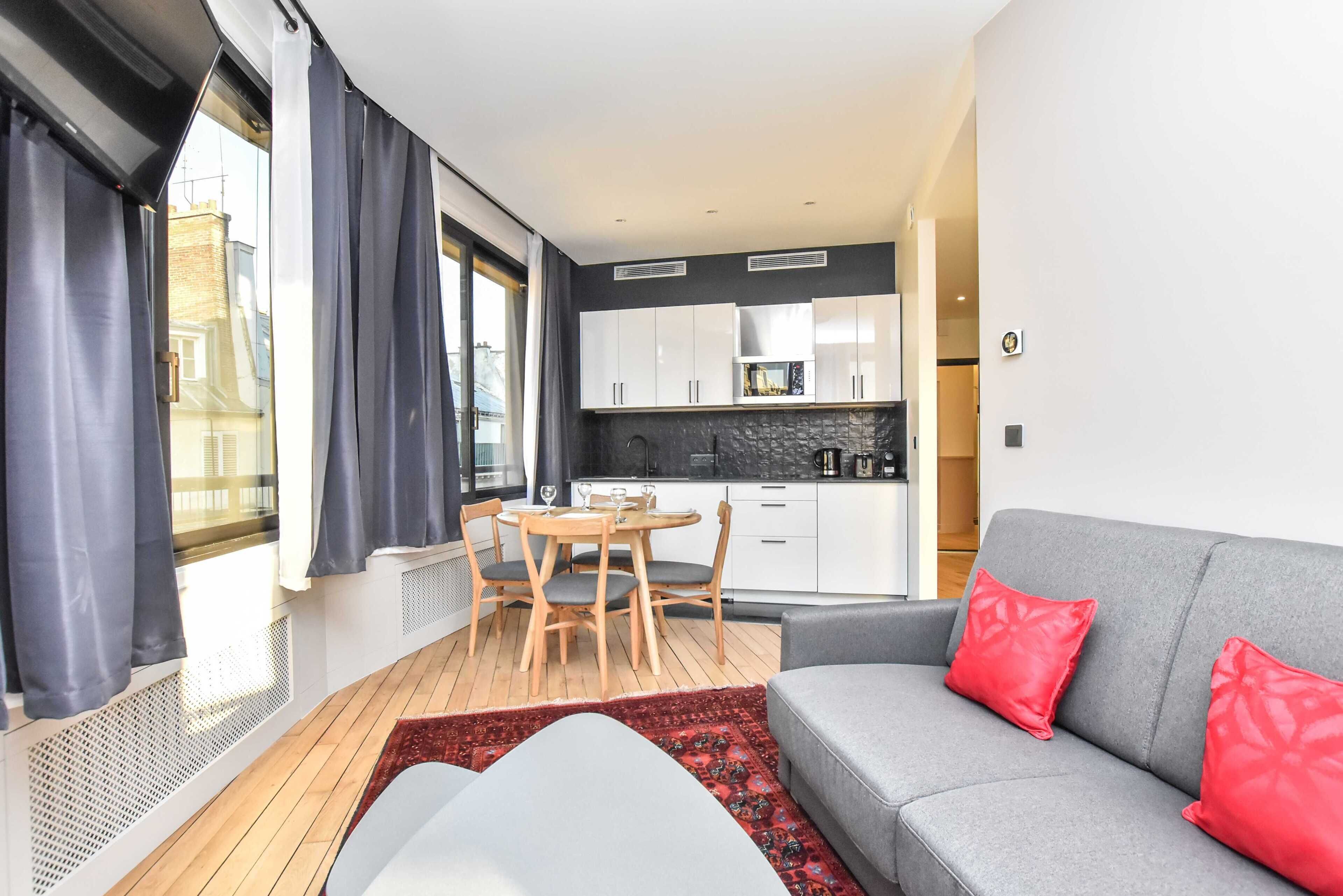 Ein modernes Wohnzimmer mit grauem Sofa und roten Kissen, ein Essbereich mit Holztisch und Stühlen sowie eine Küche mit weißen Schränken und einer schwarzen Arbeitsplatte.