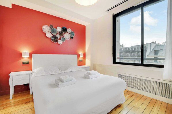 A modern hotel room with a large bed, white bedding, a red accent wall, and a city view through a large window. - Residence Paris Helder (Paris)