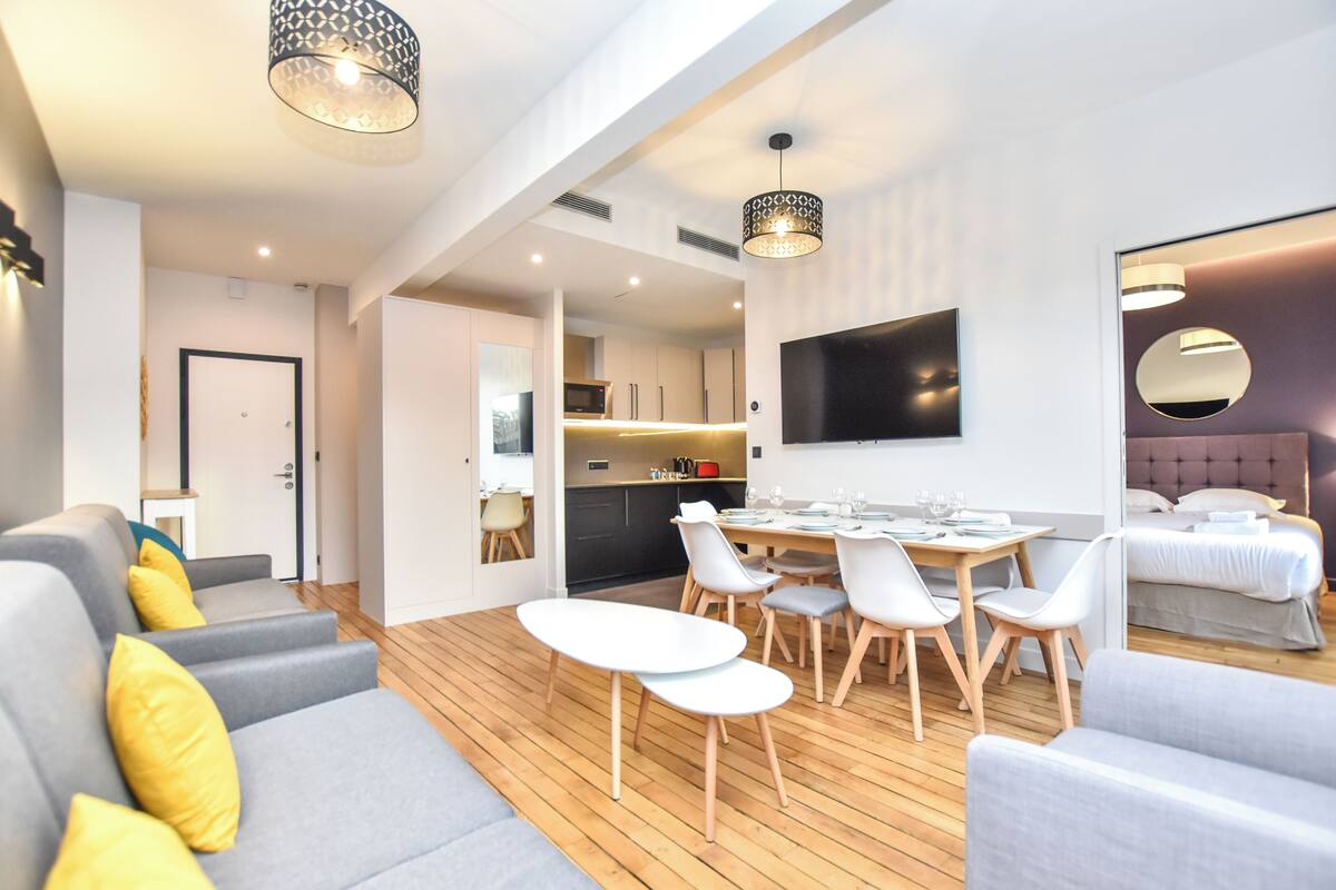 Ein modernes Wohnzimmer mit Essbereich, einer Küche im Hintergrund und einem Schlafzimmer mit großem Bett und Spiegel.