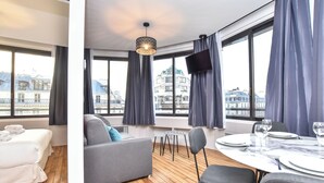 Egy modern hotelszoba, melyben fa padló, szürke színű kanapé, egy két személyre megterített étkezőasztal és nagy ablakok találhatók, melyek kilátást nyújtanak a városra.