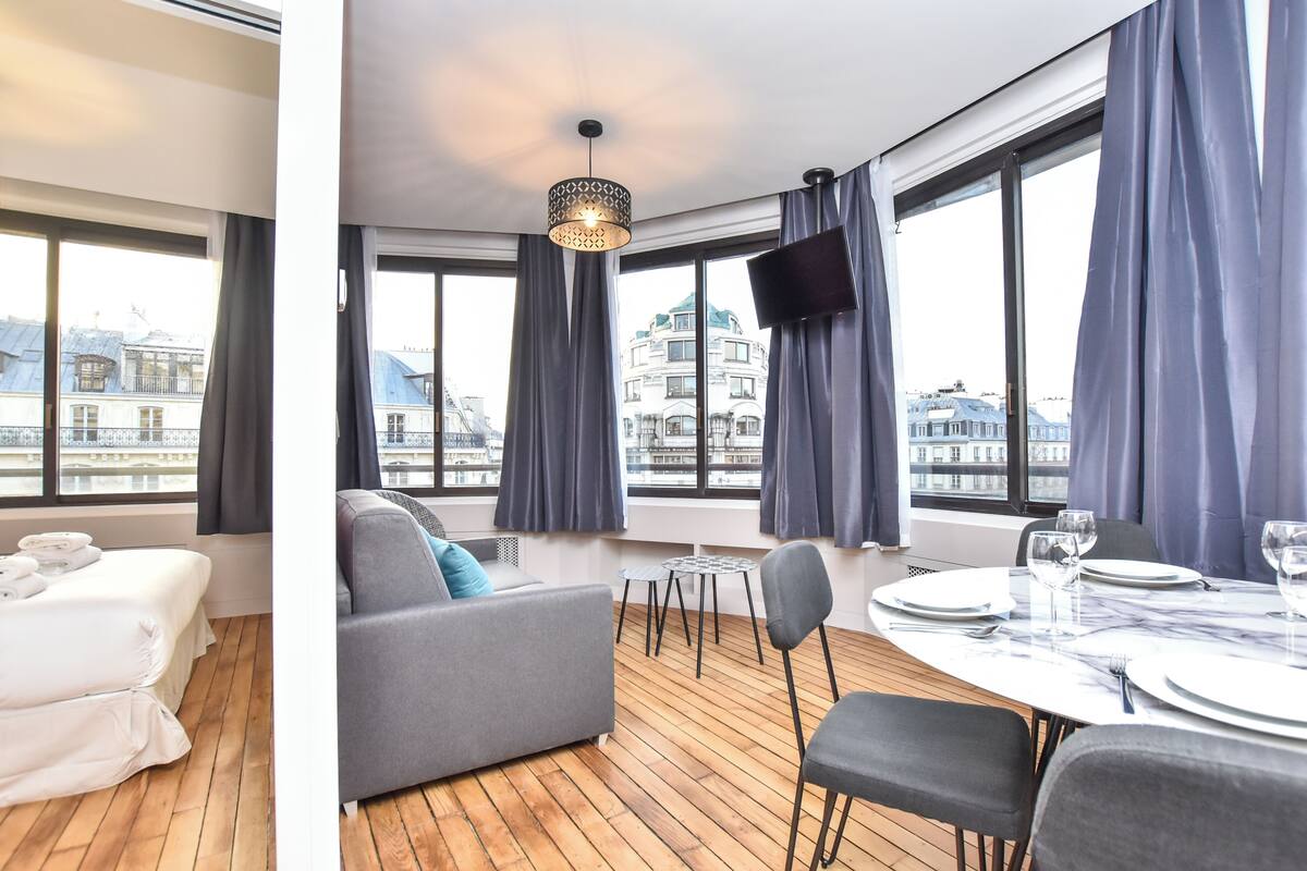 Ein modernes Hotelzimmer mit Holzboden, einem grauen Sofa, einem Esstisch für zwei Personen und großen Fenstern mit Blick auf die Stadt.