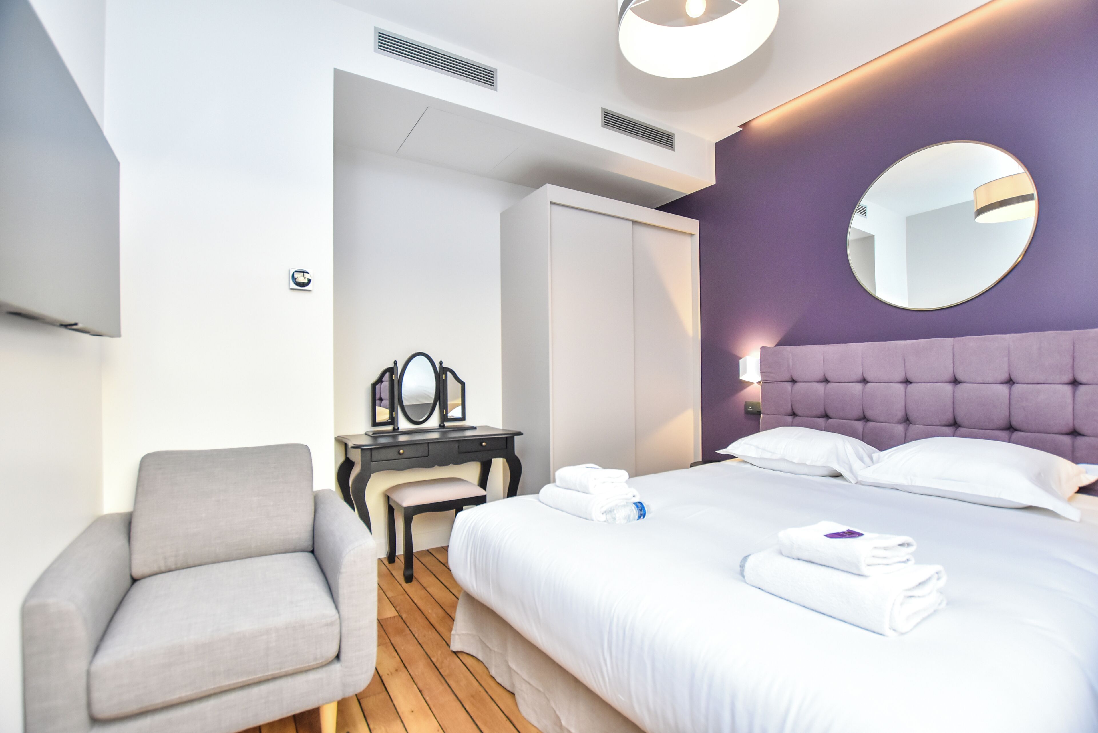 A modern hotel room with a bed, a chair, a desk, and a mirror.