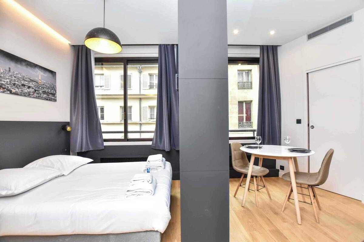 Ein modernes Hotelzimmer mit einem Bett, einem Esstisch mit Stühlen, einer Pendelleuchte und Blick auf Gebäude durch das Fenster.