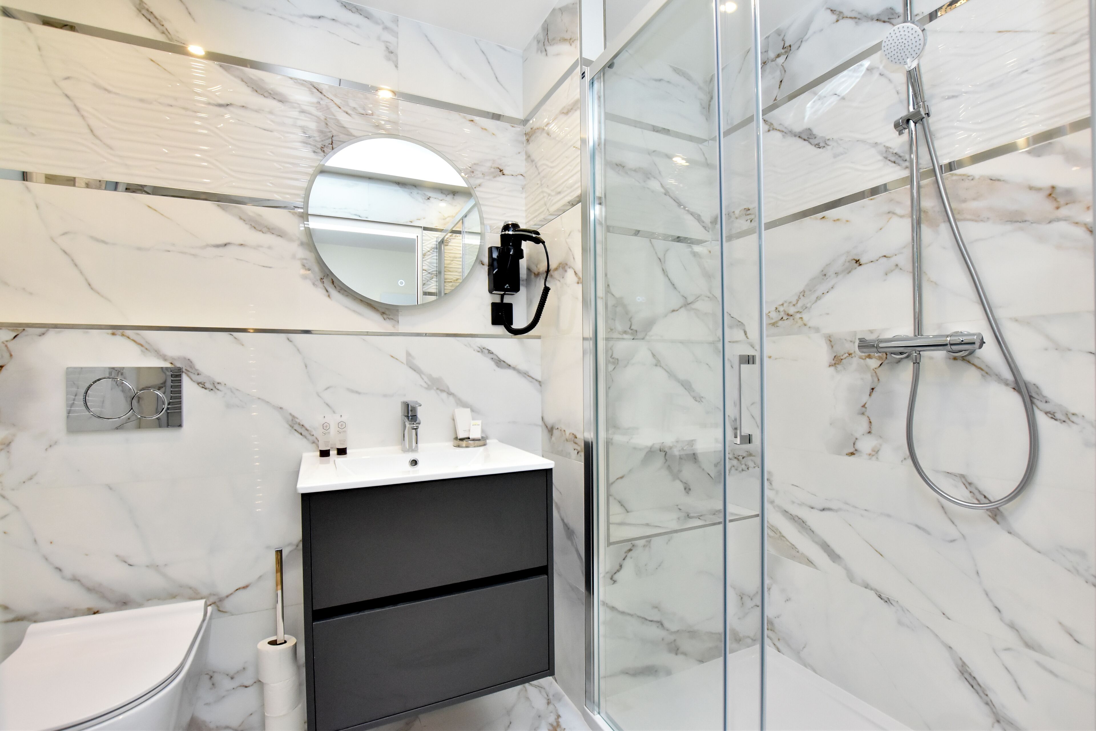 A modern bathroom with a marble shower, a glass-enclosed shower, a round mirror, and a dark-toned vanity.