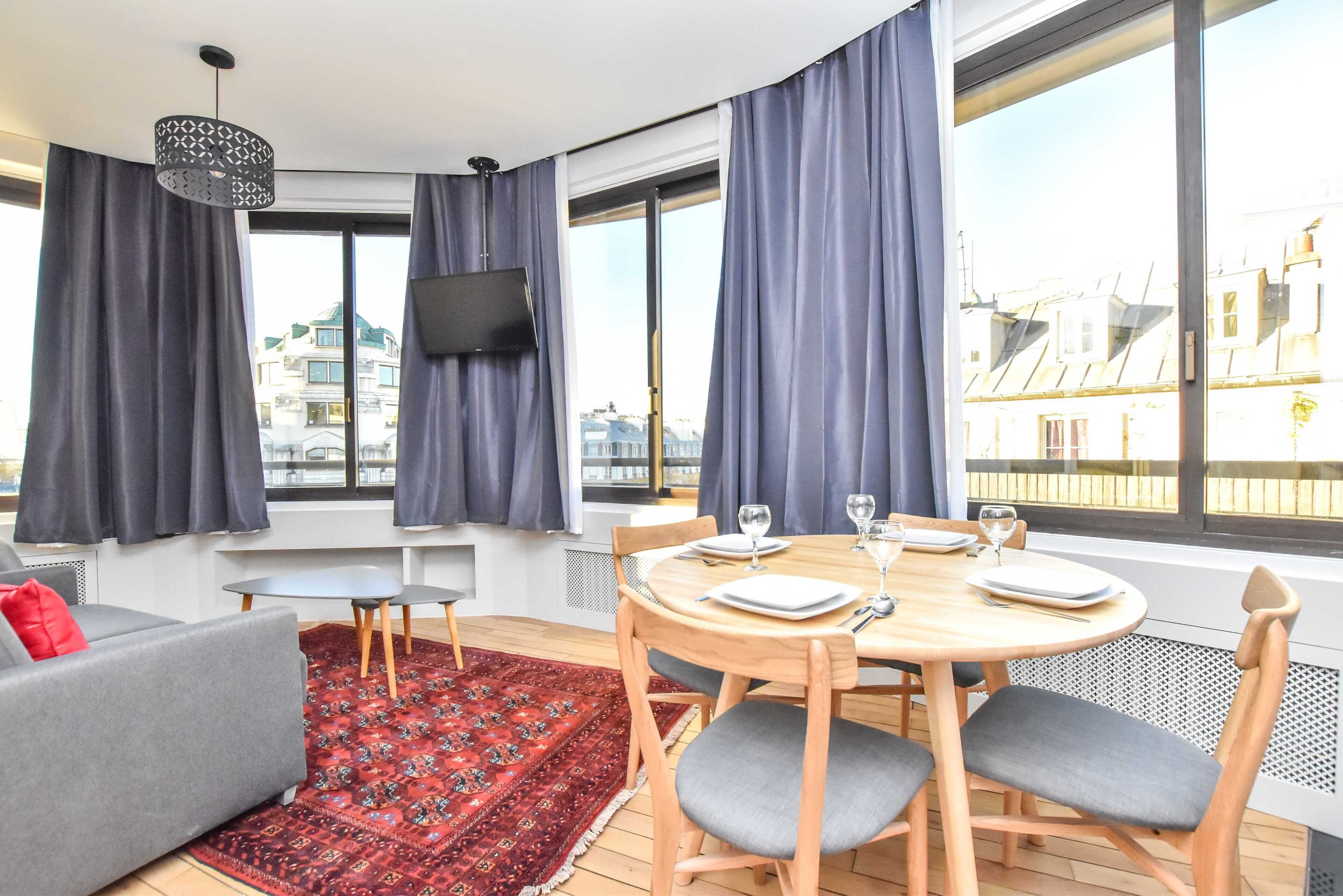 Una sala de estar moderna con un sofá, una mesa de comedor preparada para cuatro personas, un televisor montado en la pared y ventanas amplias con cortinas.
