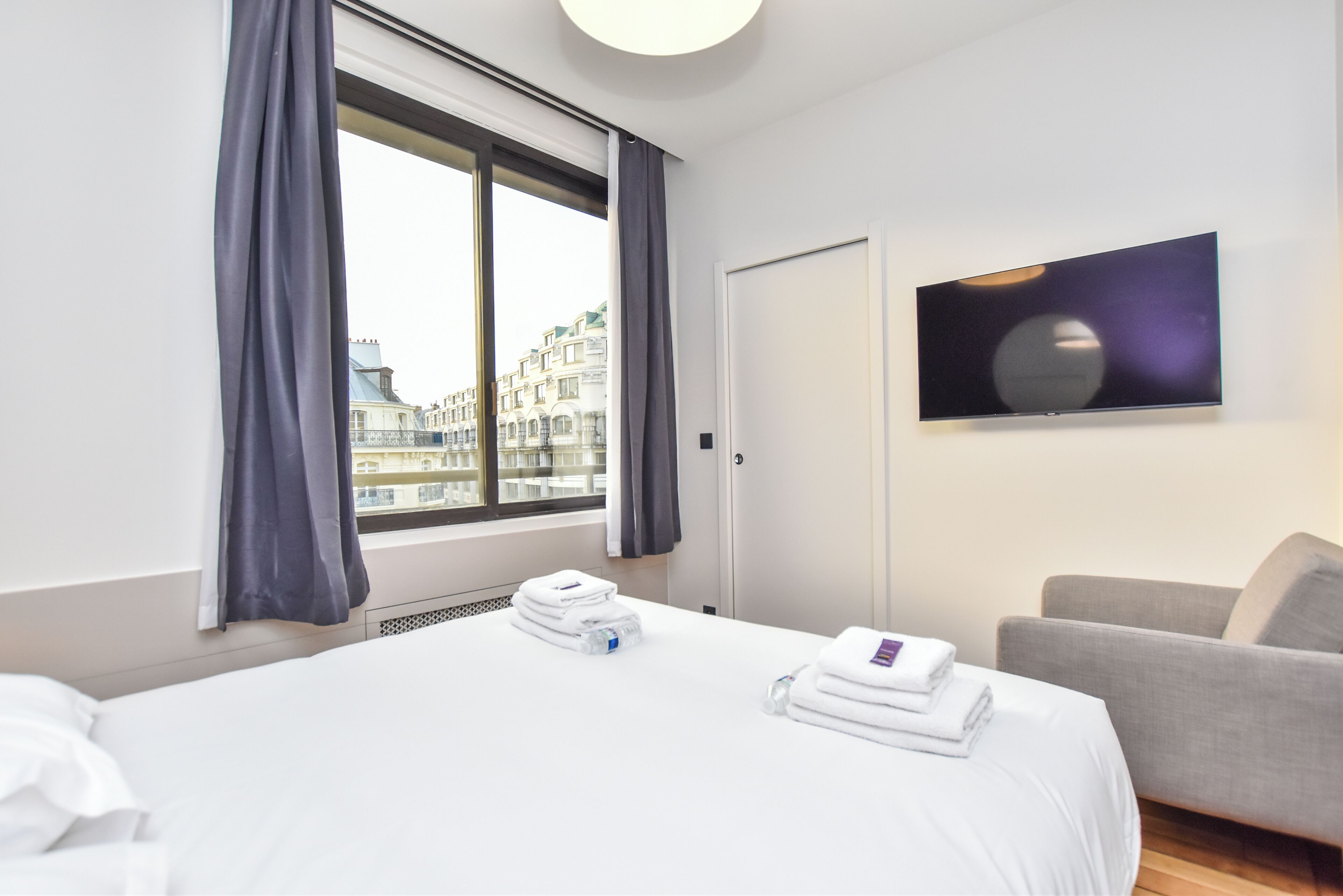 A hotel room with a bed, a TV, a grey sofa, and a window with curtains.