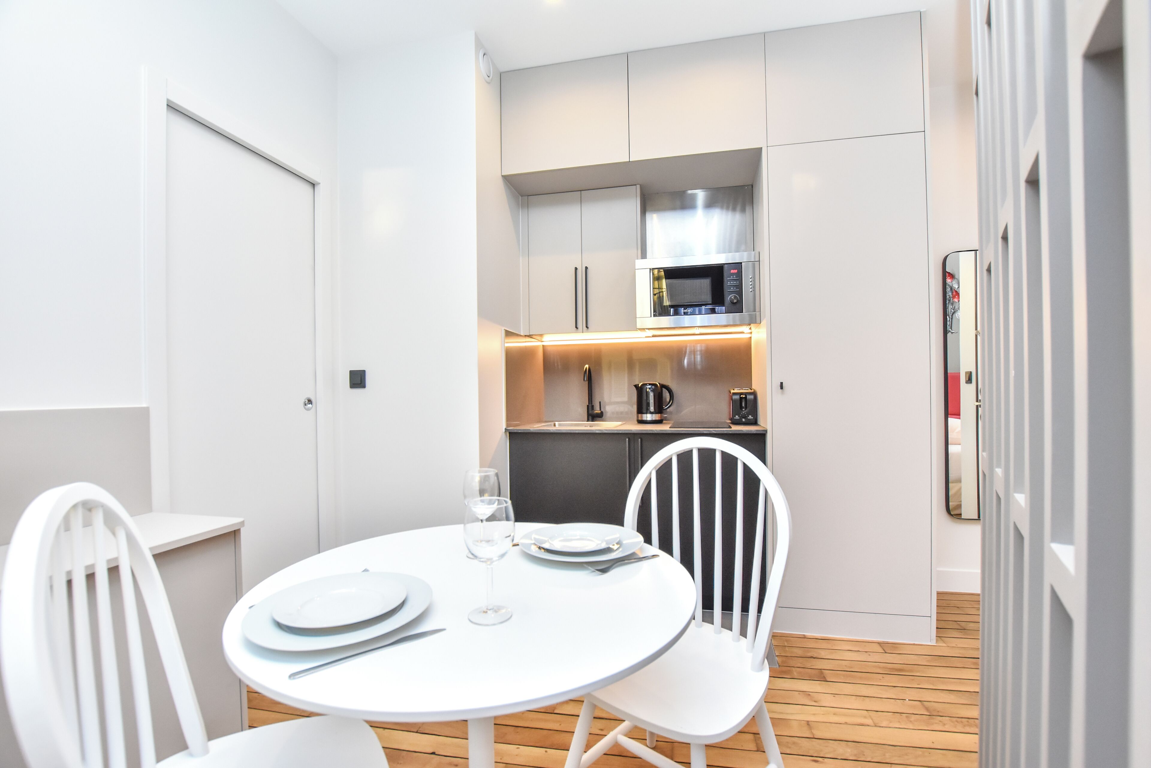A compact kitchen with a microwave, coffee maker, and kettle. There is a dining table with chairs and a glass of water.