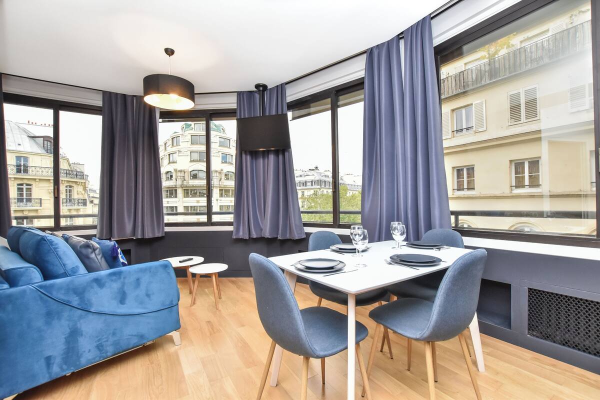 Ein moderner Essbereich mit einem weißen Tisch für vier Personen, blauen Stühlen und einem blauen Sofa. Große Fenster bieten Blick auf Gebäude und einen Balkon.