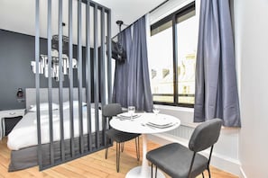 Una habitación de hotel moderna con cama, escritorio, mesa redonda con sillas y un gran ventanal con cortinas.