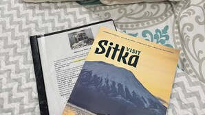 Un panier contenant des en-cas et une brochure intitulée « Sitka » est posé sur un lit recouvert d’une literie à motifs.