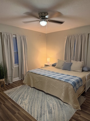 Un dormitorio con cama, cortinas, ventilador de techo, lámpara de noche y alfombra.