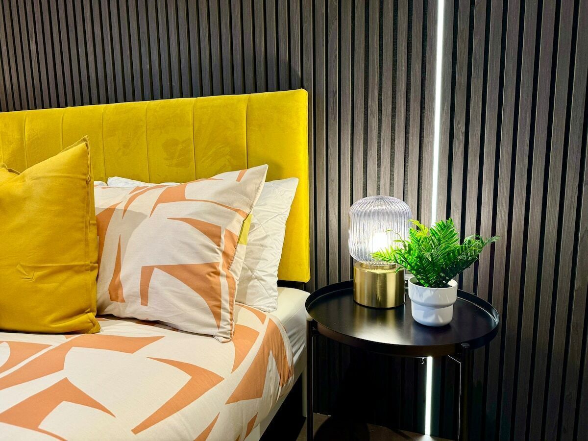 Una cama con cabecera amarilla, ropa de cama blanca y naranja con estampado, una mesita de noche redonda negra con una planta y una lámpara decorativa.