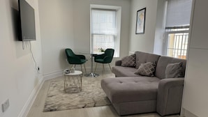 Ein modernes Wohnzimmer mit einem grauen Sofa, zwei grünen Sesseln, einem kleinen runden Tisch und einem an der Wand befestigten Fernseher.