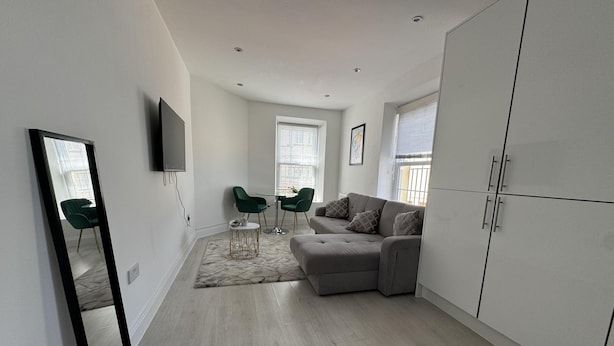 Ein modernes, minimalistisches Wohnzimmer mit einem grauen Sofa, grünen Sesseln, einem kleinen runden Tisch und einem großen Kleiderschrank.