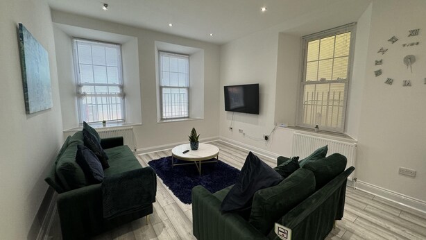 A modern living room with two green sofas, a round coffee table, and a flat-screen TV mounted on the wall.
