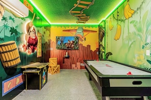 Una habitación con mesa de billar, dardos y una pantalla grande mostrando un videojuego.