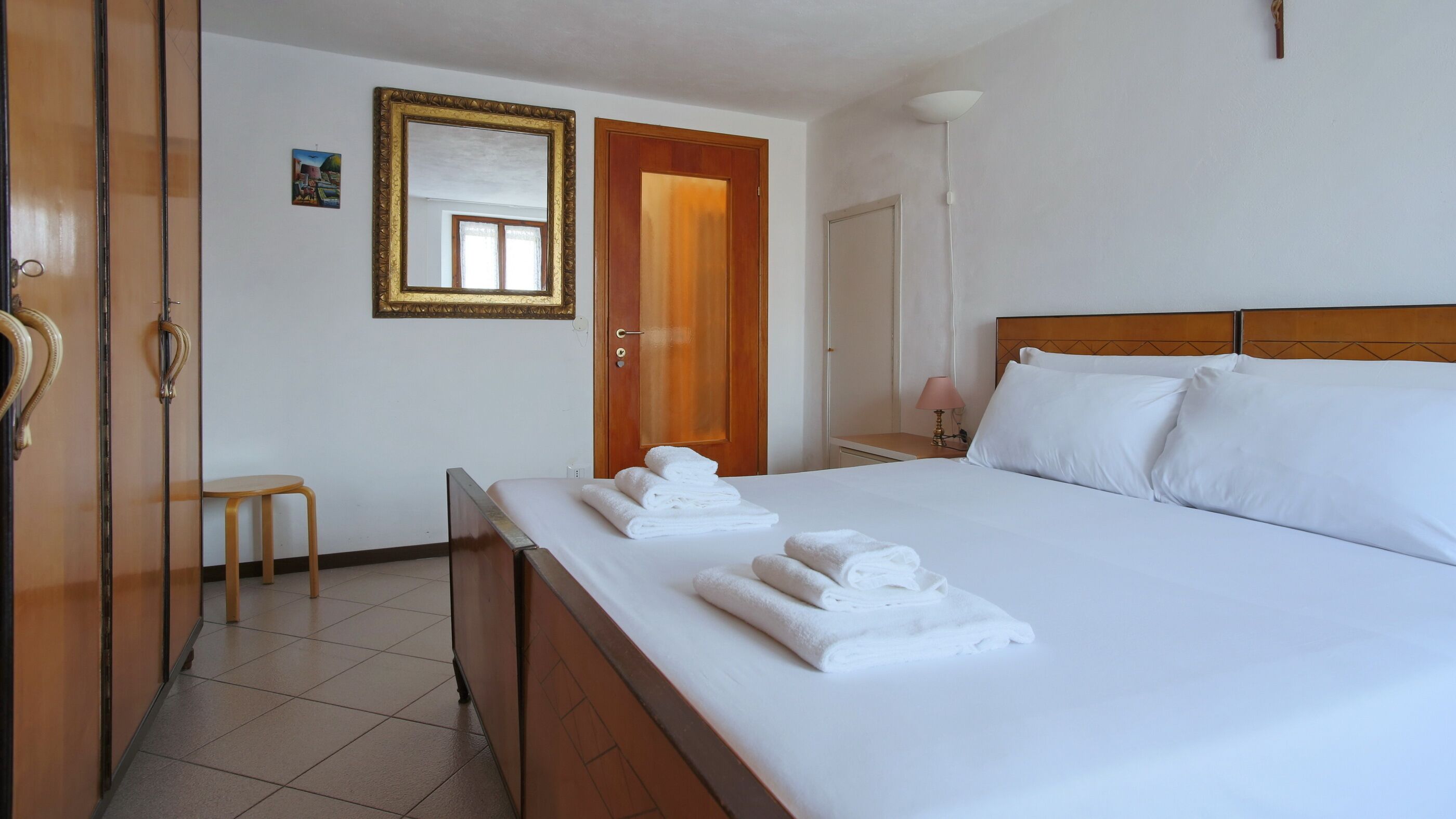 A hotel room with a bed, a wooden headboard, a nightstand with a lamp, a small table, and a door with a mirror.