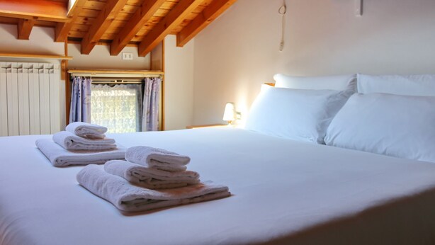 Una cama bien hecha con sábanas y toallas blancas, techo de madera y una ventana con cortinas.