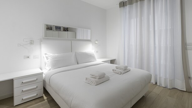 Une chambre d’hôtel moderne avec un lit bien fait, une table de chevet, une armoire intégrée et une grande fenêtre agrémentée de rideaux légers.