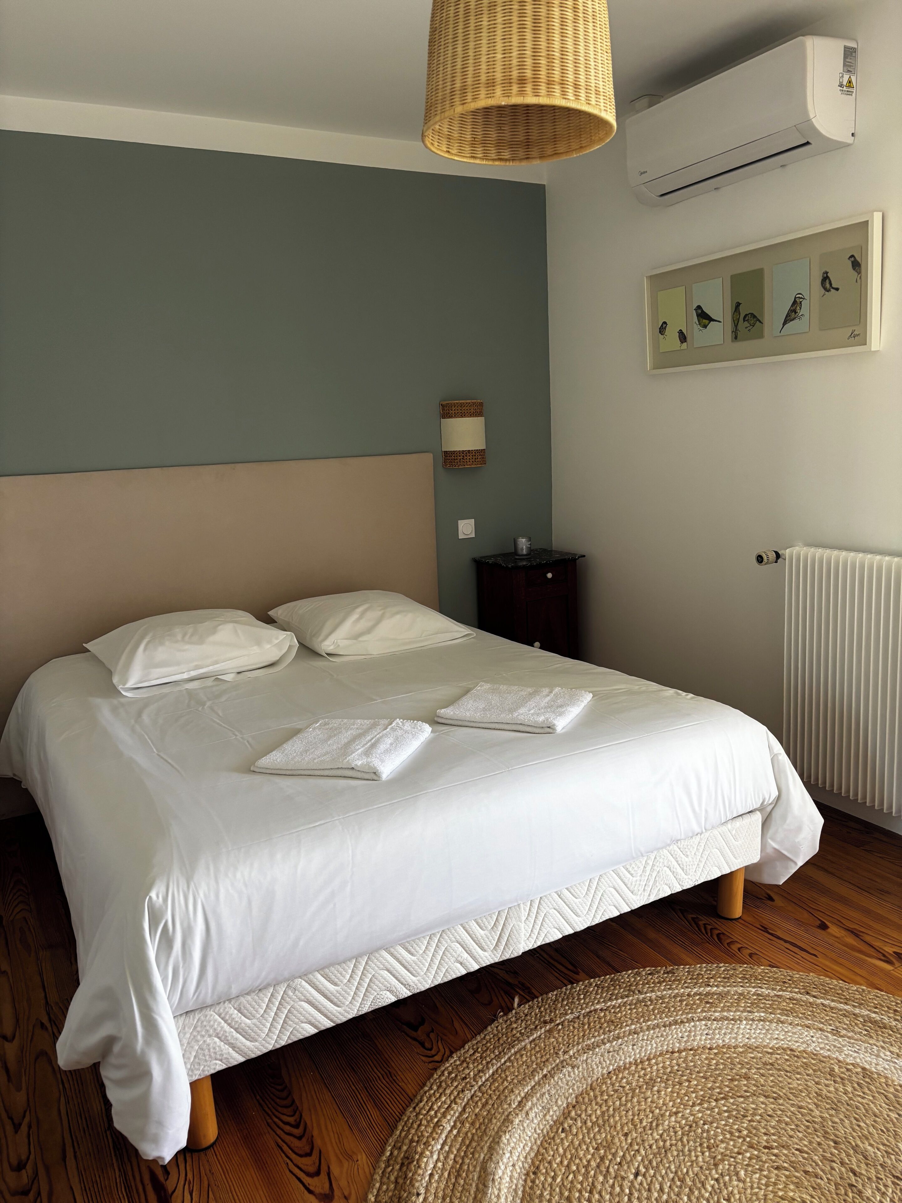 Un lit bien fait, avec des oreillers et des serviettes pliées, une lampe en osier, un climatiseur, un tableau représentant un oiseau encadré, une petite table de chevet et un radiateur.