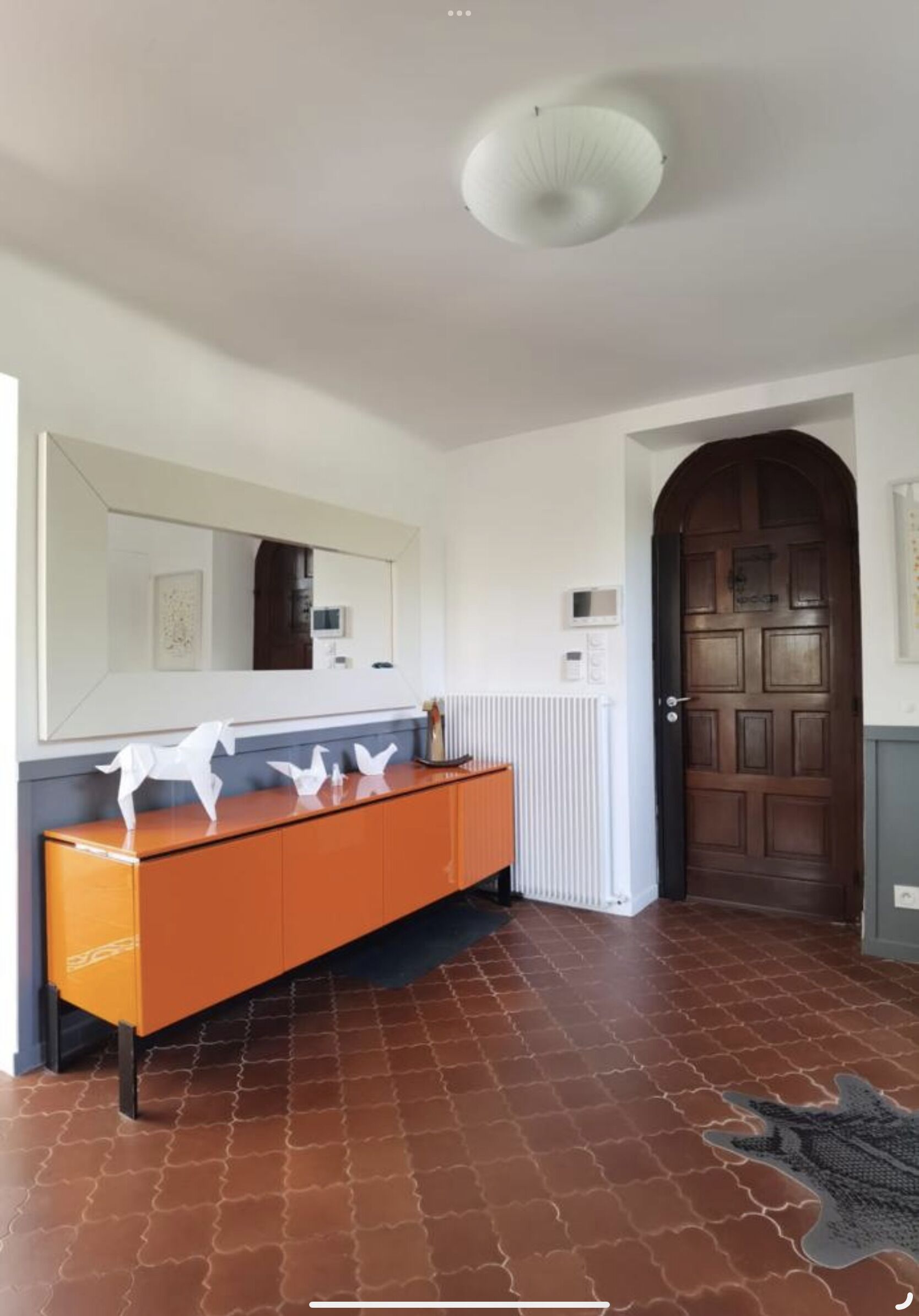 Een gang met een bruine betegelde vloer, een houten deur, een witte radiator en een spiegel boven een houten console met witte paardenbeeldjes.