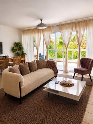 A living room with a sofa, a coffee table, and a chair. There is a dining table with chairs and a plant. The room has large windows with curtains. - Beach Condo 3 mins walking distance from the beach, Amazing Pool 2BR Gated Comm. (Punta Cana)