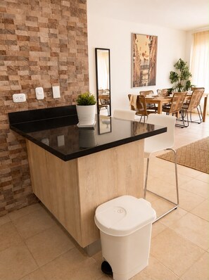A modern kitchen with a black countertop, a white trash bin, and a brick wall. - Beach Condo 3 mins walking distance from the beach, Amazing Pool 2BR Gated Comm. (Punta Cana)