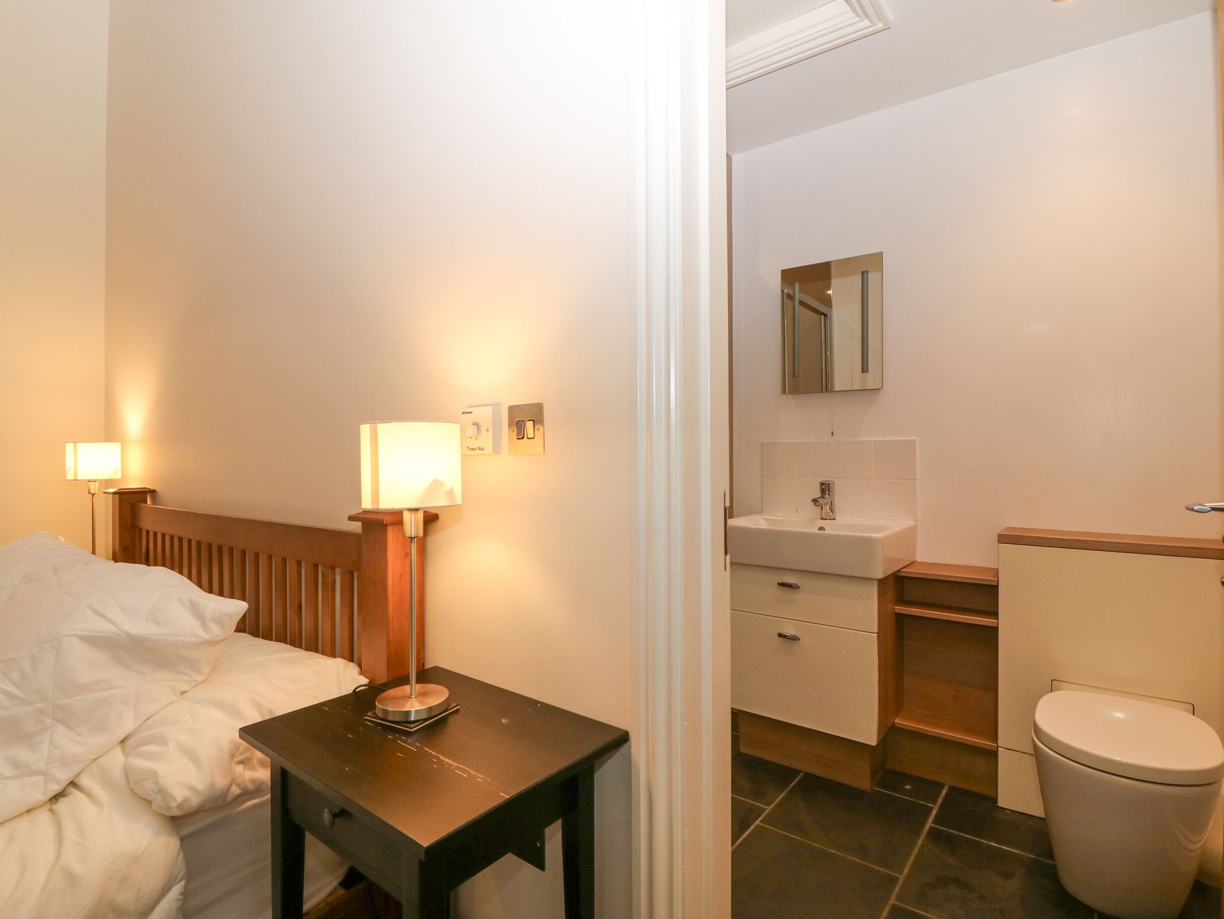 Une chambre d’hôtel comprenant un lit, une table de chevet avec une lampe, une petite table, une salle de bain avec un lavabo, un miroir et des toilettes.